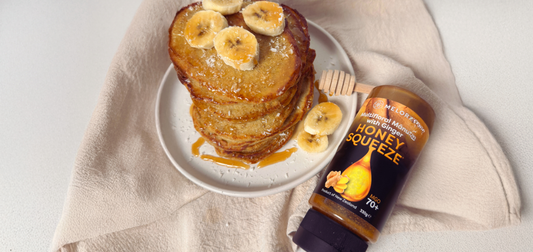 A stack of banana pancakes topped with fresh banana slices, served alongside a bottle of Melora Multifloral Mānuka Honey with Ginger.